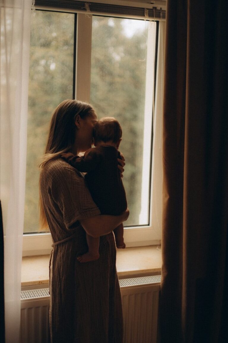 A mother holds her baby close beside a window in dim warm light