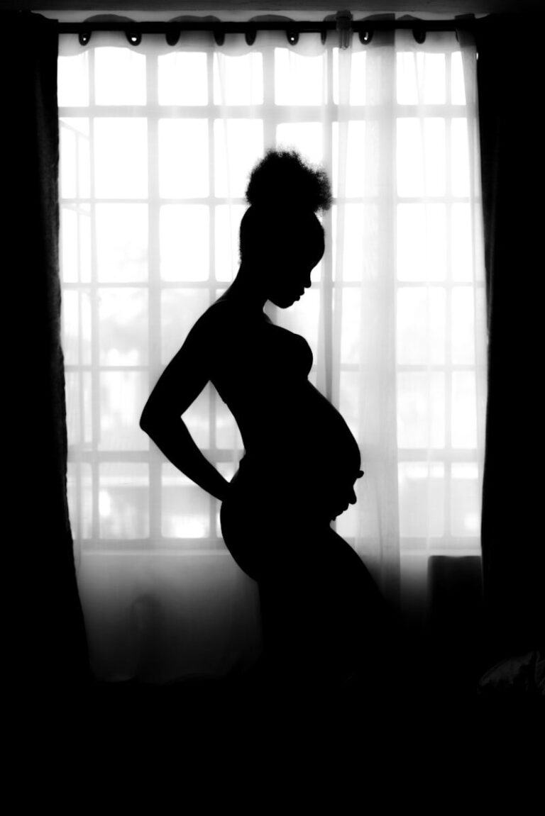 A pregnant woman stands in profile, silhouetted against a bright window with sheer curtains.