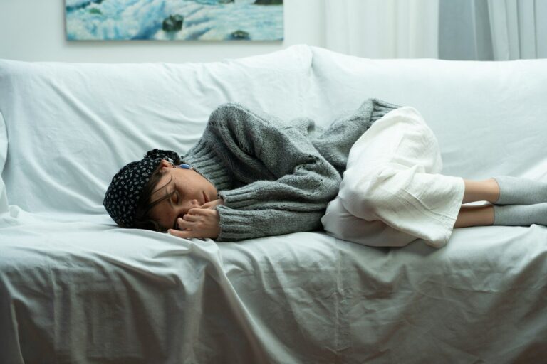 A woman in a cozy gray sweater lies curled up resting on a white couch in soft daylight.