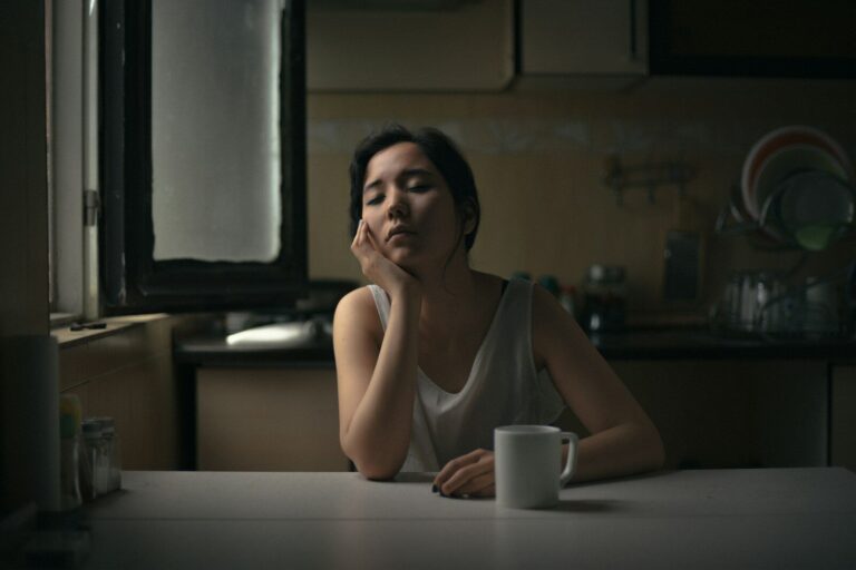 A weary woman rests her chin on her hand at a kitchen table beside a mug of coffee