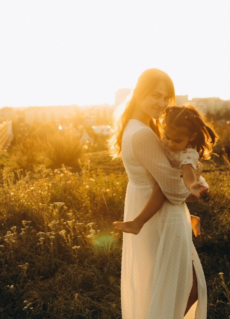 A mother in a white dress holds her young daughter close in a sunlit field at golden hour.