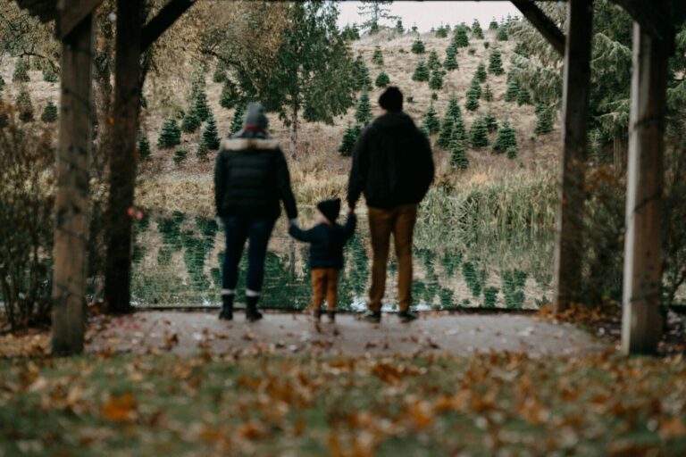 Two parents and their small child stand hand in hand looking out at a quiet lake framed by trees.