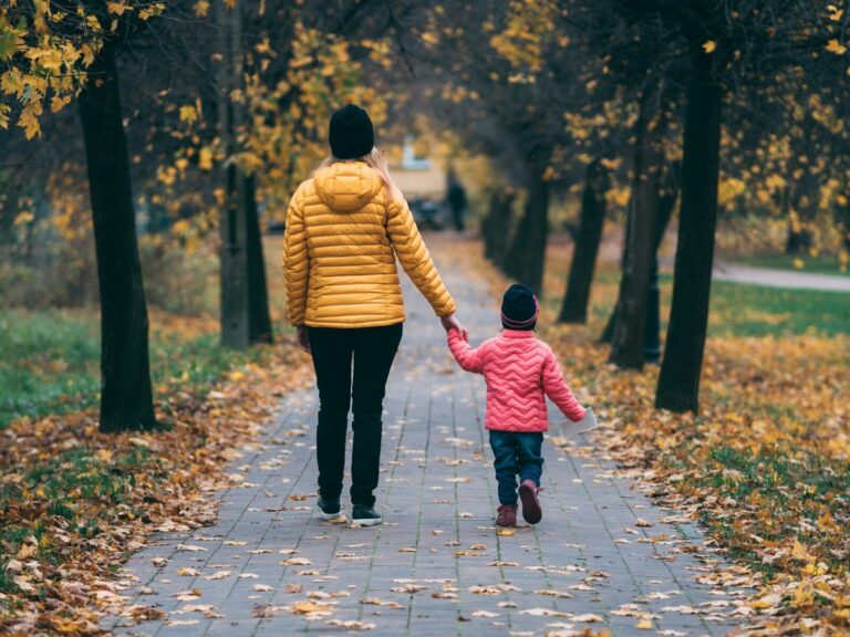 A mother and her young child walk hand in hand down a tree-lined path scattered with autumn leaves.