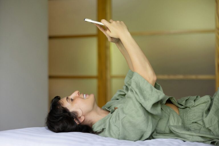 A woman lies in bed holding her phone above her face in soft morning light